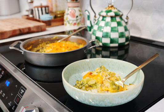 Bowl of chicken broccoli divan served over rice on a stovetop, skillet of casserole and green-checkered teapot in background