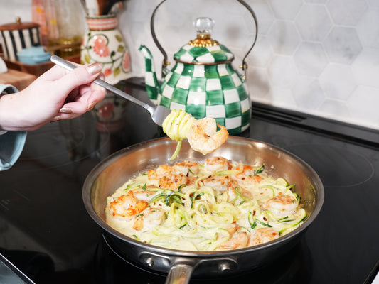 Hand holding a fork lifting a shrimp-wrapped zucchini noodle from a skillet of creamy garlic sauce on a stovetop