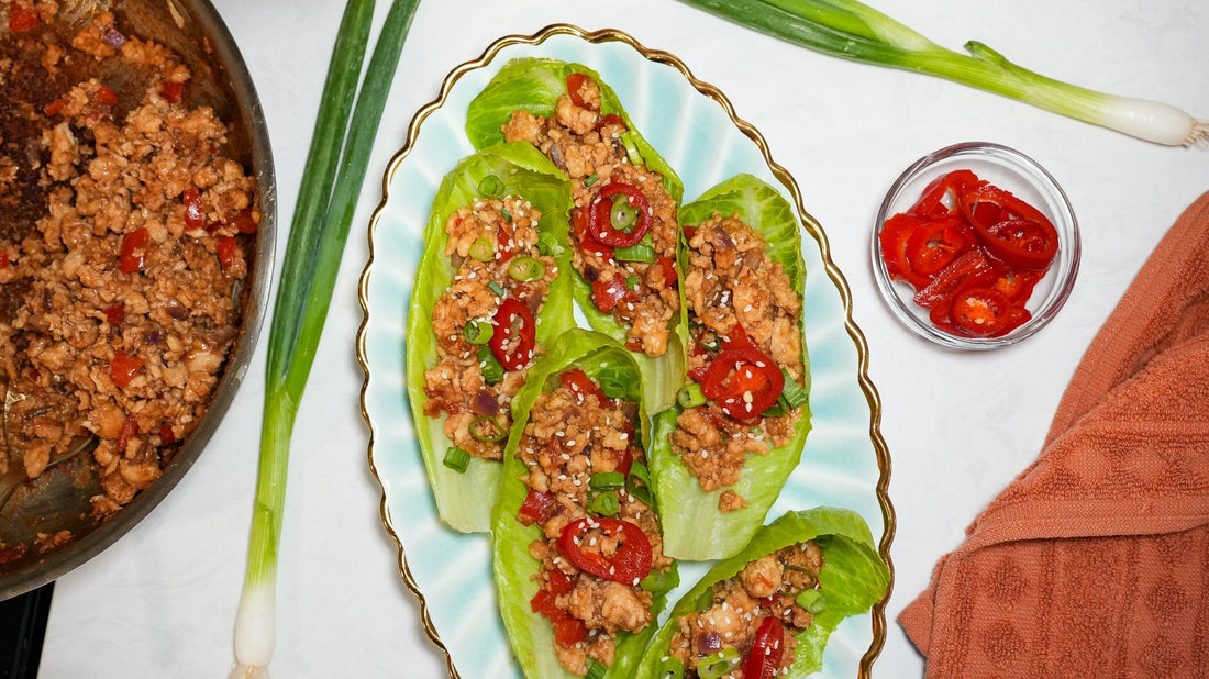 Three Asian chicken lettuce wraps topped with sliced red chili, scallions, and sesame seeds on a scalloped plate