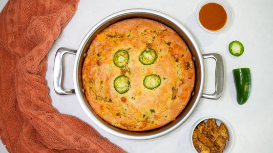 Top view of barbecue pork-stuffed cornbread in a round stainless steel pan topped with jalapeño slices, with towel and dipping sauce on a white surface