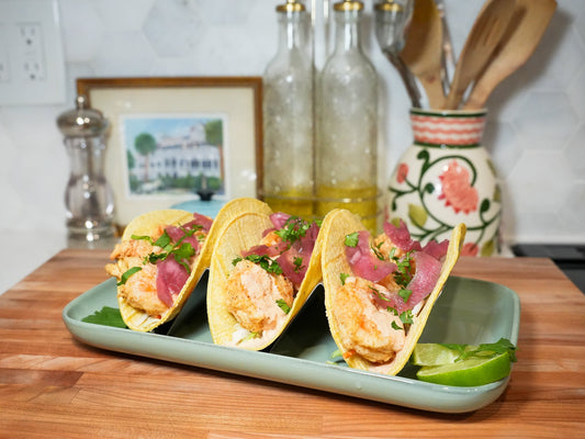 Three shrimp tacos topped with pickled red onions and cilantro on a green plate on a wooden counter in a kitchen setting