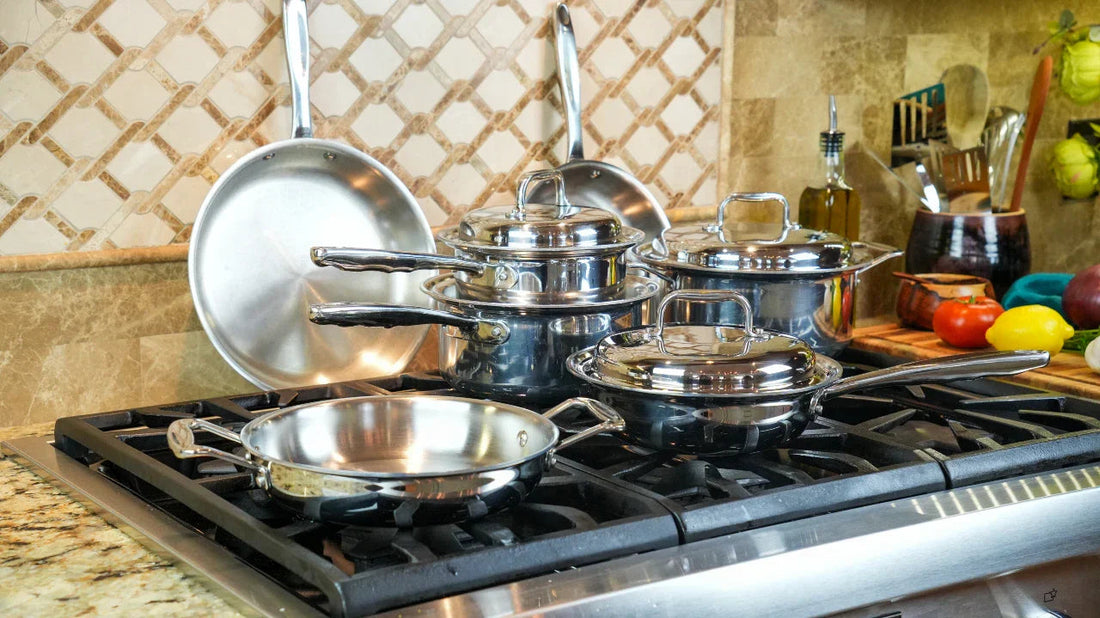 Angled view of stainless steel cookware set (frying pans and saucepans with lids) showcasing polished mirror finish on a gas stovetop in a tiled kitchen
