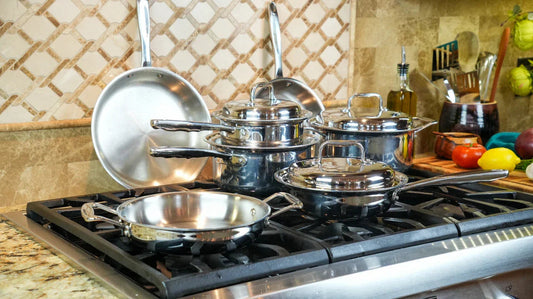 Angled view of stainless steel cookware set (frying pans and saucepans with lids) showcasing polished mirror finish on a gas stovetop in a tiled kitchen