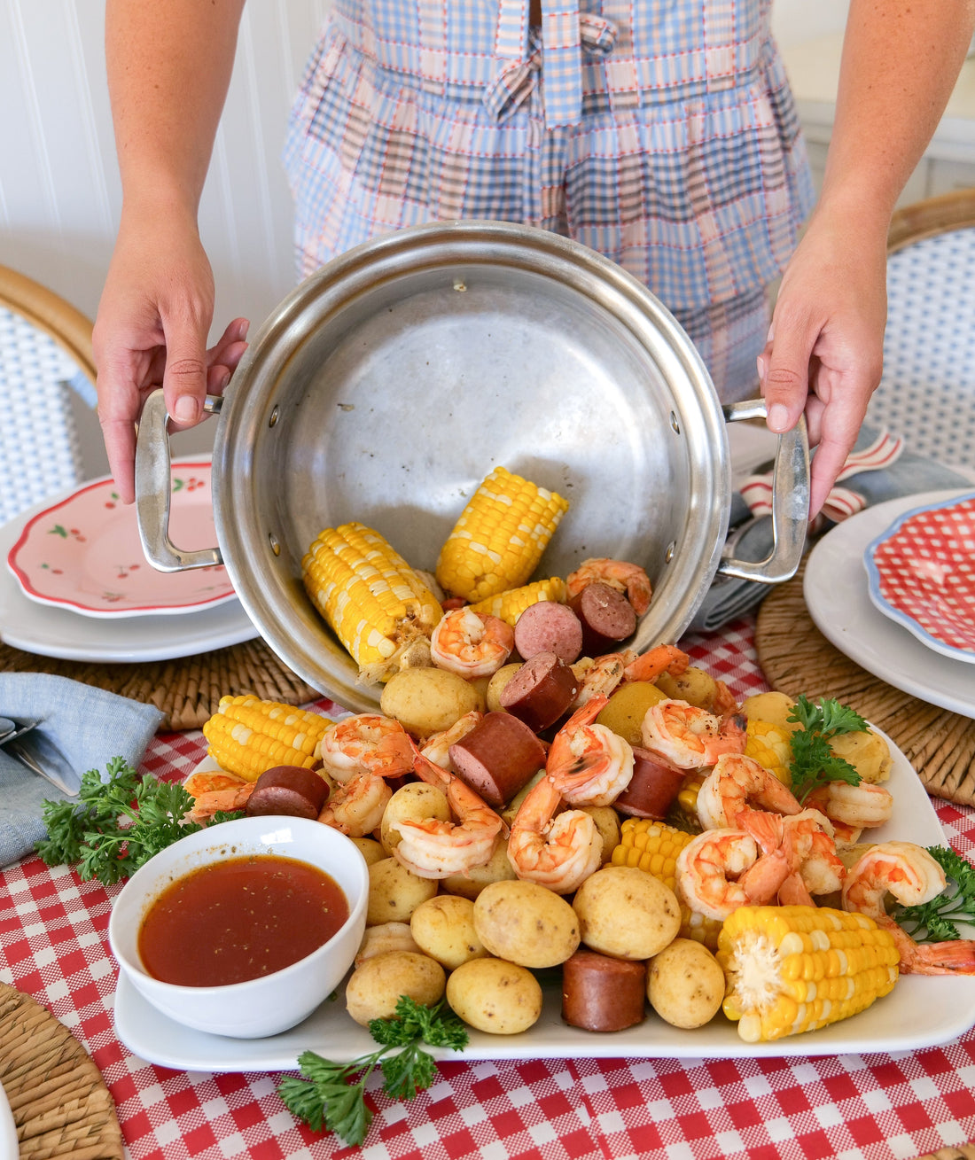 Person pouring shrimp boil with corn, potatoes, shrimp, and sausage from a stainless-steel pot onto a platter over a red-checkered tablecloth