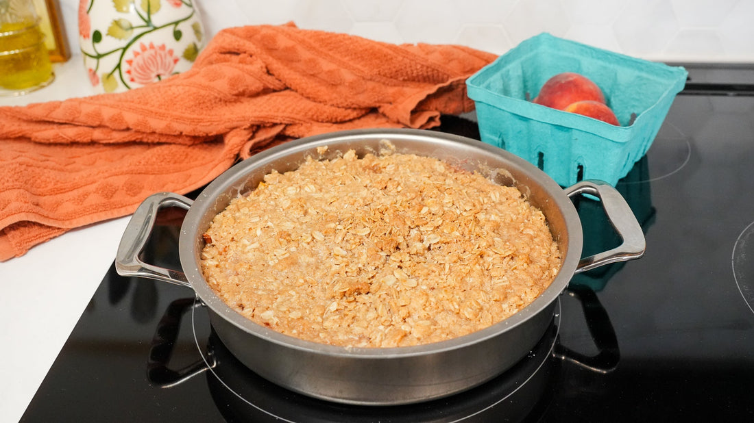 Peach crisp in a round skillet on a black stovetop with a teal carton of peaches and a folded orange kitchen towel