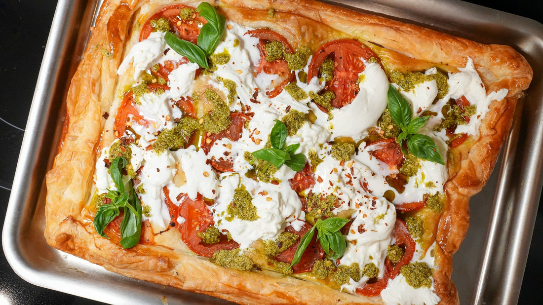 Overhead view of a tomato tart topped with torn burrata, dollops of pesto, and fresh basil on a metal baking sheet