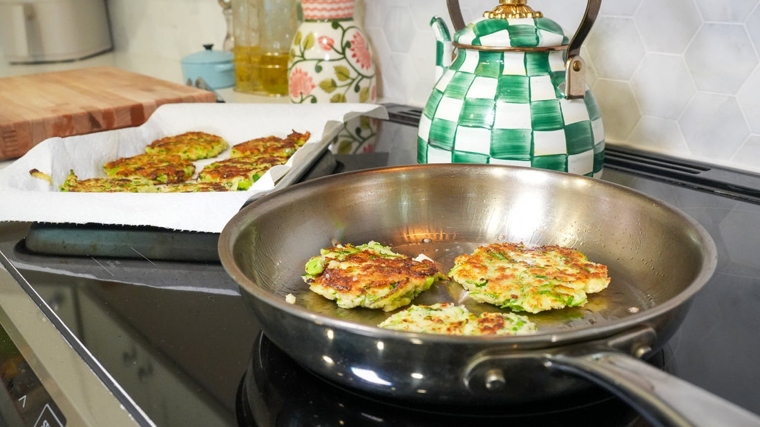 Zucchini fritters frying in a stainless steel skillet on a stovetop, tray of cooked fritters on paper towel in the background