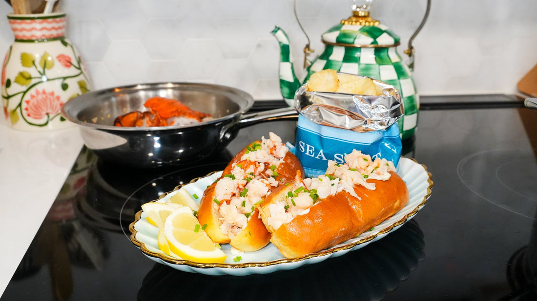 Two lobster rolls topped with chopped lobster and chives on a decorative plate with lemon wedges, a bag of chips, and a pot on a stovetop