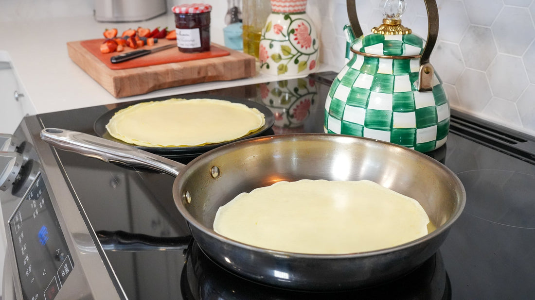 Crepes cooking in a stainless steel skillet on a glass-top stove with a green checkered kettle behind