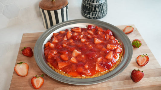 Cream cheese strawberry pie in a round pan on a wooden cutting board, topped with glazed strawberry chunks and surrounded by halved strawberries