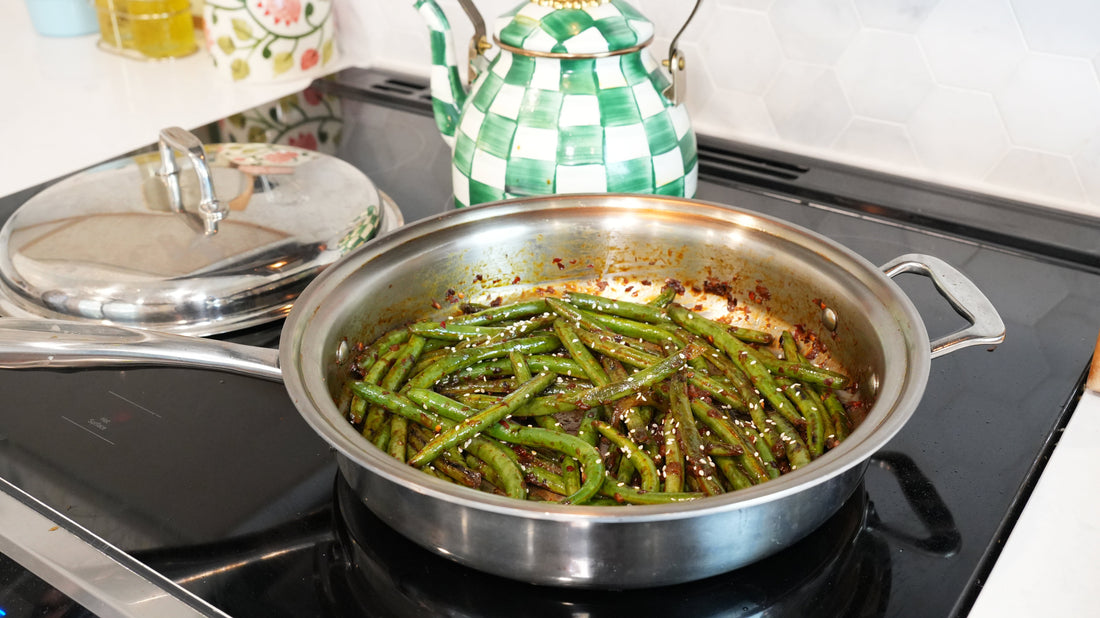 Sichuan-style green beans sautéing in a stainless-steel pan on a stovetop, garnished with sesame seeds