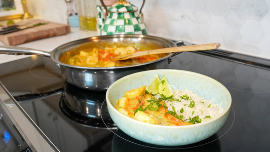 Bowl of coconut curry shrimp with white rice and lime wedges on stovetop, stainless-steel pan and wooden spoon in background