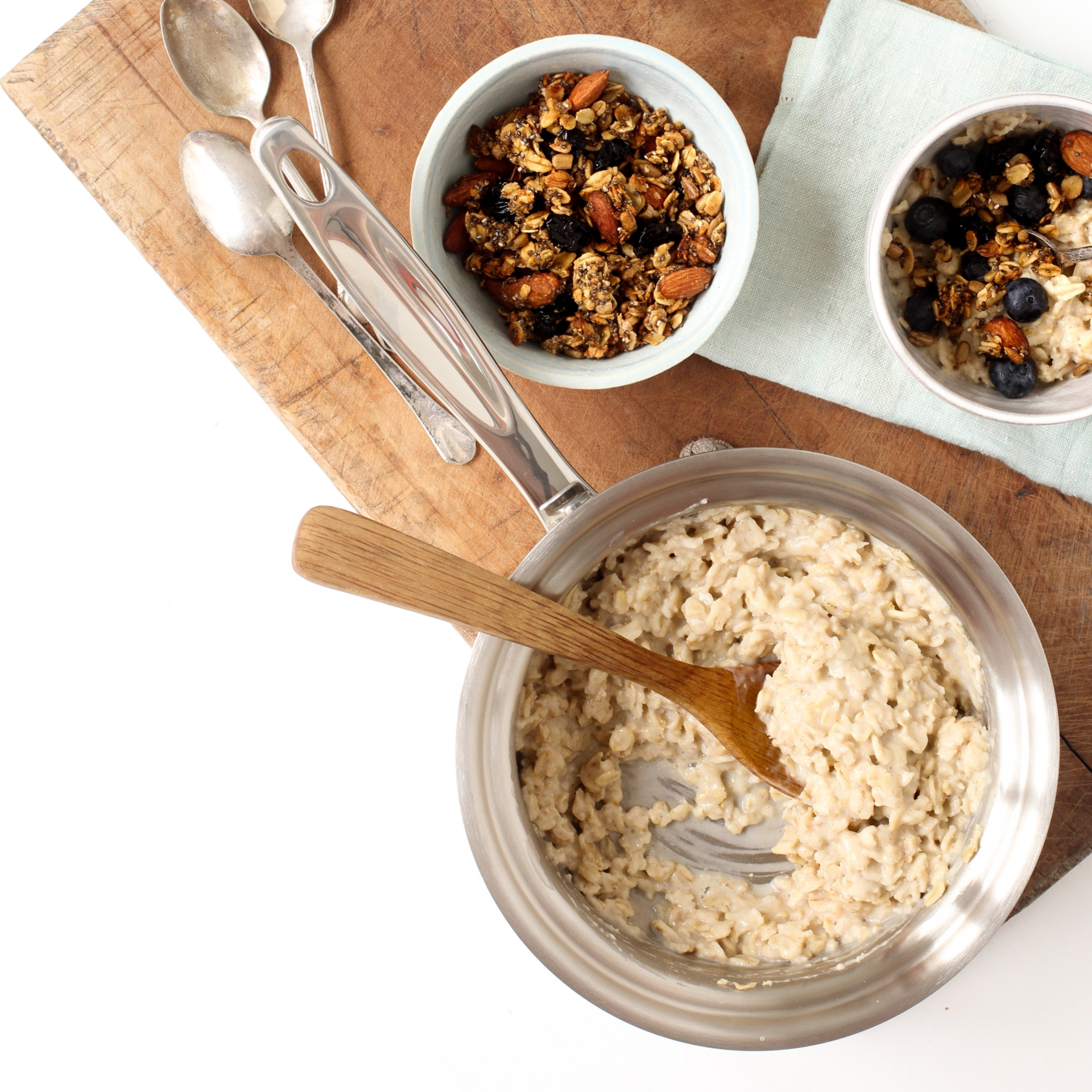 A saucepan with oatmeal and bowls of granola on a wooden cutting board.