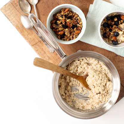 A saucepan with oatmeal and bowls of granola on a wooden cutting board.