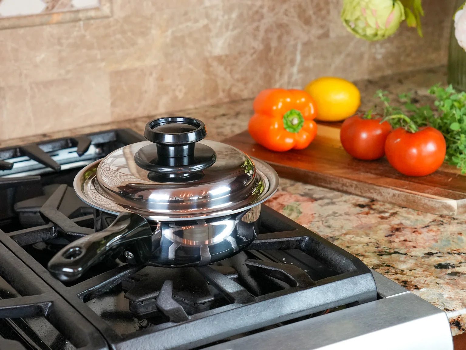 A stainless steel saucepan with a clear glass lid and black handle sits on a black gas stovetop in a kitchen. A wooden cutting board with tomatoes, a yellow pepper, and herbs is visible in the background against a beige stone backsplash.