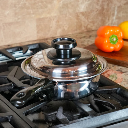 A stainless steel saucepan with a matching glass lid and black handle, sitting on a black gas stovetop in a kitchen setting with a tiled backsplash and colorful bell peppers in the background.