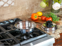 A stainless steel saucepan sits on a gas stovetop in a kitchen setting, with a matching lid placed beside it on a granite countertop. Fresh vegetables, including tomatoes, a yellow bell pepper, and herbs, are arranged on a wooden cutting board, and a vase with flowers is visible in the background.