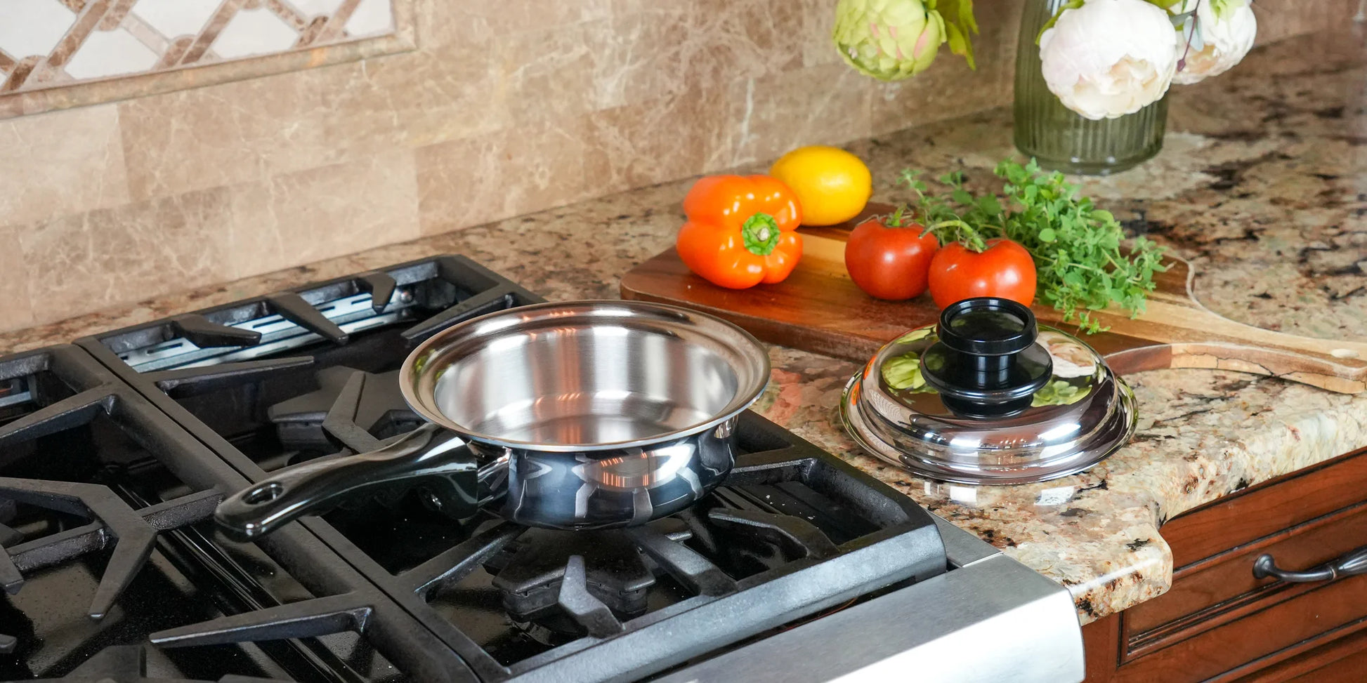 A stainless steel saucepan sits on a black gas stovetop burner in a kitchen setting, with a matching lid nearby on a granite countertop and fresh vegetables in the background.