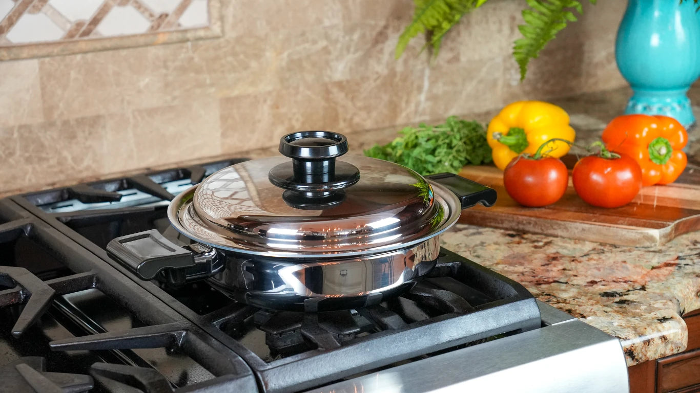 A stainless steel pot with a clear glass lid sits on a gas stovetop in a kitchen setting, with fresh vegetables and a blue vase in the background.