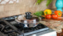 A stainless steel pot with a clear glass lid sits on a gas stovetop in a kitchen setting, with fresh vegetables and a blue vase in the background.