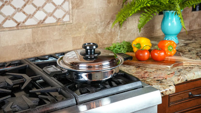 A stainless steel saucepan with a glass lid sits on a gas stovetop in a kitchen setting, with fresh tomatoes, peppers, and a blue vase on the countertop nearby.