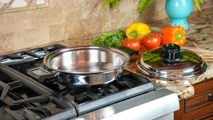 A stainless steel sauté pan sits on a gas stovetop, with its matching lid resting on a granite kitchen counter. Fresh vegetables, including tomatoes, peppers, and greens, are arranged in the background against a beige tile backsplash.