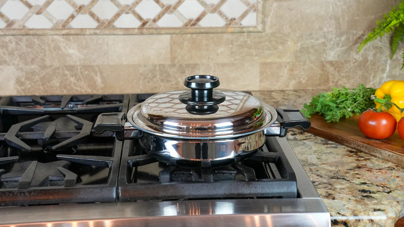 A stainless steel pot with a glass lid sits on a gas stovetop in a kitchen setting, with a marble tile backsplash and fresh vegetables on a cutting board in the background.