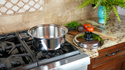 A stainless steel pot with a matching lid sits on a gas stovetop in a kitchen setting, with a granite countertop, vegetables, and a blue vase in the background.