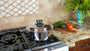 A stainless steel pot with a glass lid sits on a gas stovetop in a kitchen setting with a tiled backsplash, granite countertop, and fresh vegetables nearby.
