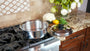 A stainless steel pot with a matching lid sits on a gas stovetop in a kitchen setting, with a granite countertop, flowers, and vegetables in the background.