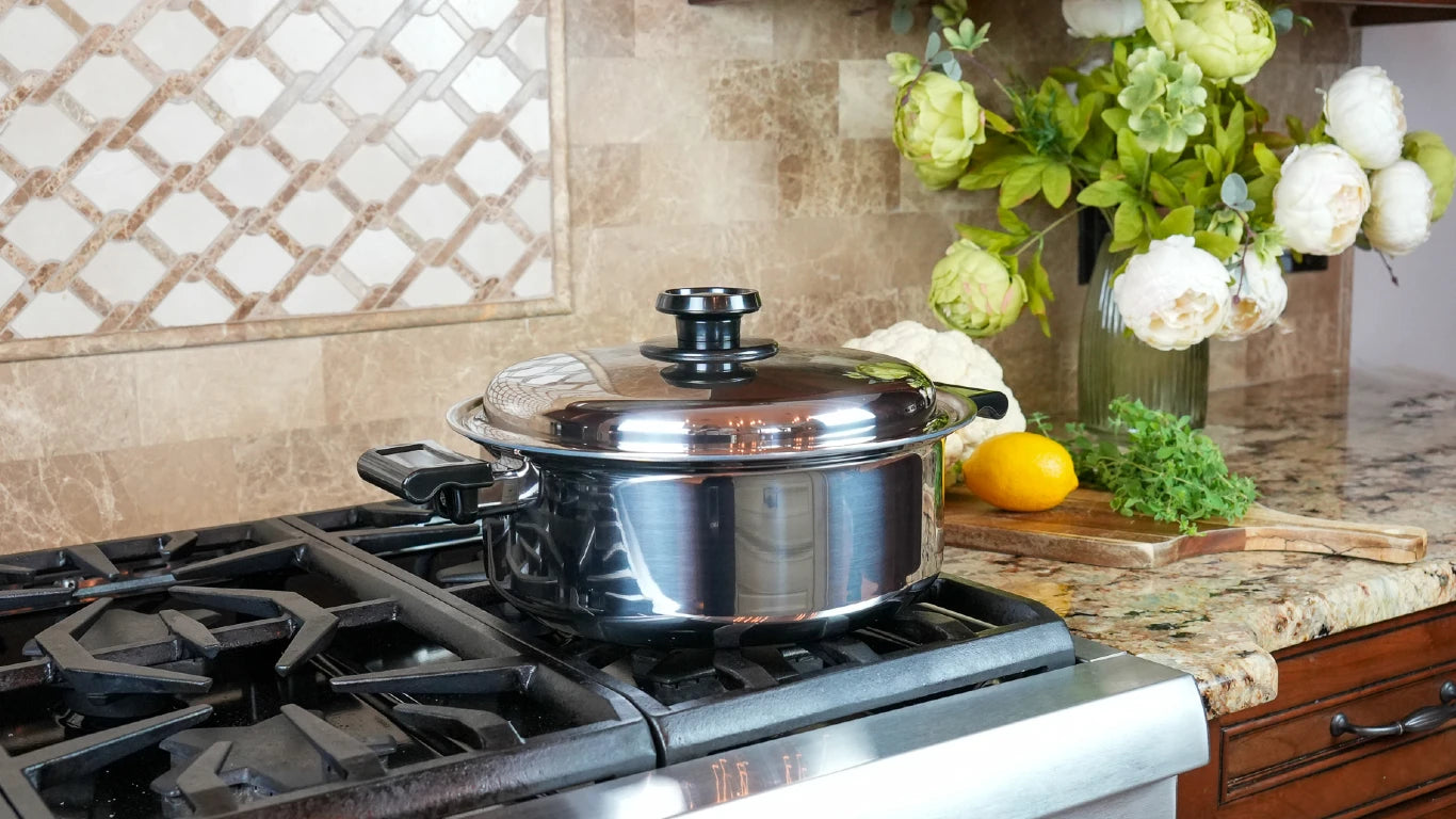 A stainless steel stockpot with a glass lid sits on a gas stovetop in a kitchen setting, with a tiled backsplash, flowers, and a lemon on the counter in the background.
