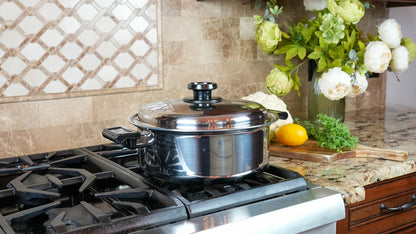 A stainless steel stockpot with a glass lid sits on a gas stovetop in a kitchen setting, with a tiled backsplash, flowers, and a lemon on the counter in the background.
