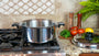 A large stainless steel stockpot with black handles sits on a gas stovetop in a kitchen setting. A matching lid rests on the granite countertop beside tomatoes, parsley, a lemon, and wooden spices, with a tiled backsplash behind.