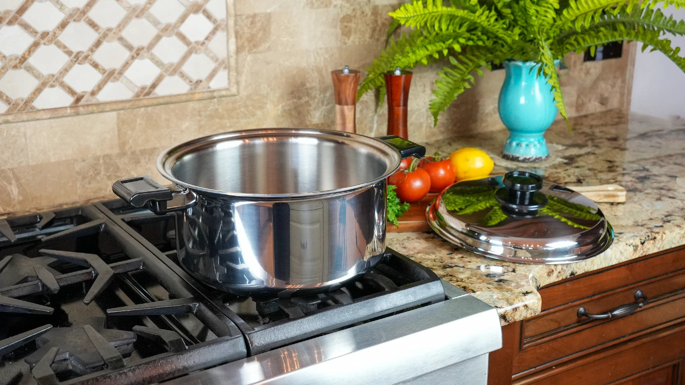 A large stainless steel stockpot sits on a gas stovetop in a kitchen setting, with its matching lid placed on the granite countertop beside tomatoes, lemons, and a blue vase.