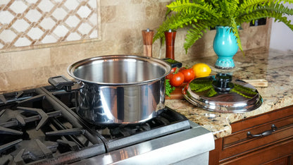 A large stainless steel stockpot sits on a gas stovetop in a kitchen setting, with its matching lid placed on the granite countertop beside tomatoes, lemons, and a blue vase.