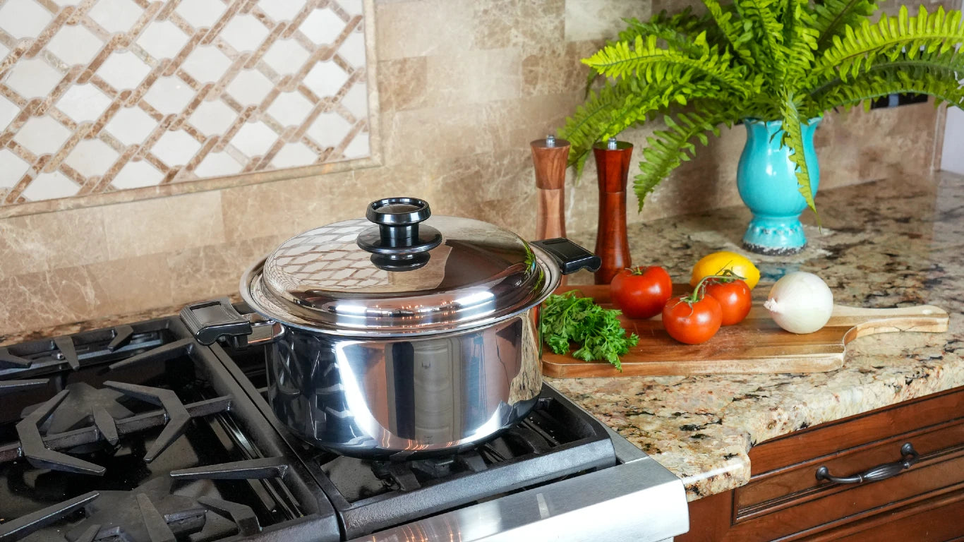 A stainless steel stockpot with a lid sits on a gas stovetop in a kitchen setting, with a cutting board, tomatoes, onion, and a vase of greenery on the granite countertop.