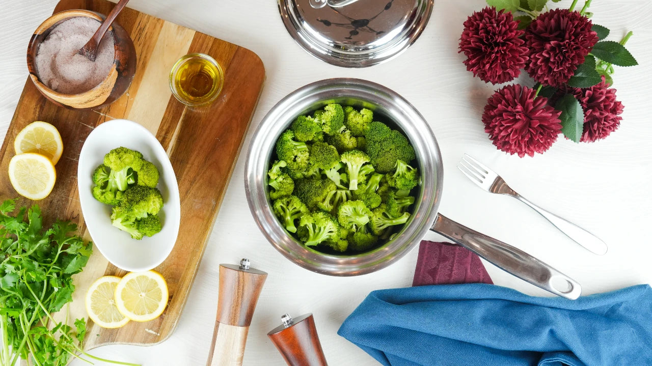 Broccoli in a bowl on a white surface with lemons, herbs, and a cutting board.