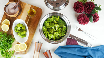Broccoli in a bowl on a white surface with lemons, herbs, and a cutting board.