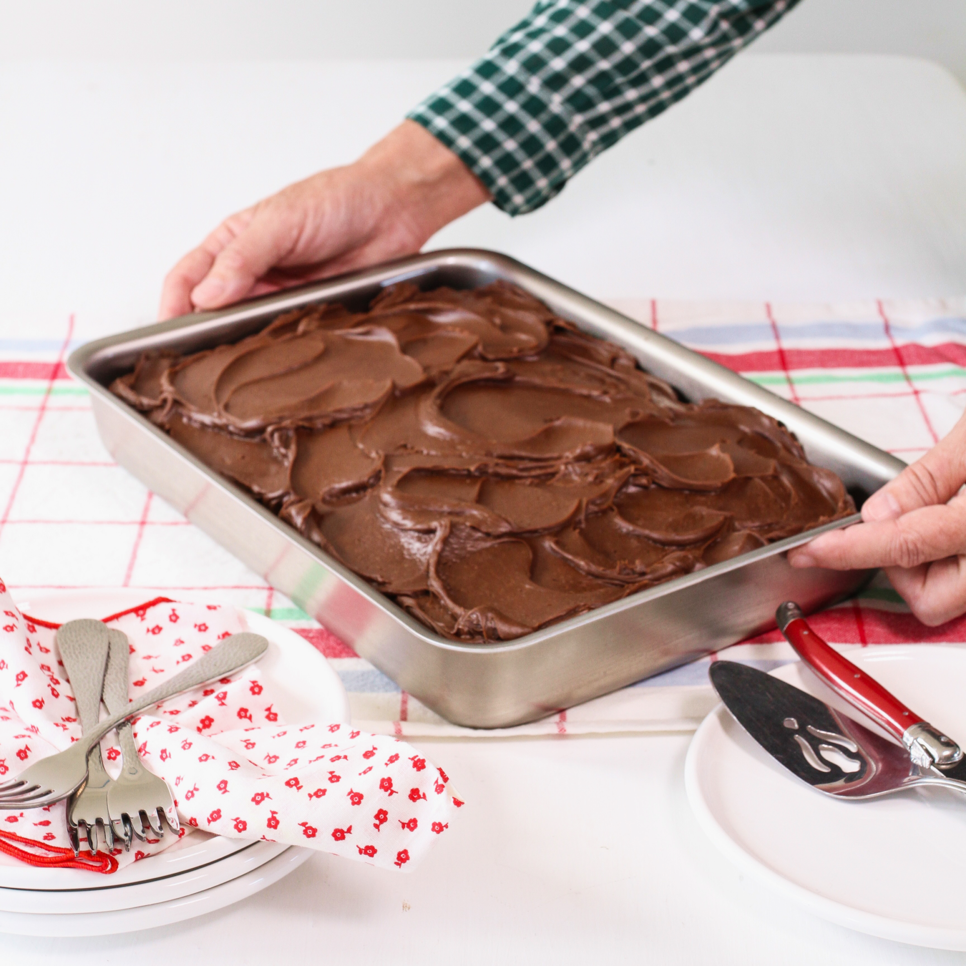 A person holding a brownie pan filled with chocolate frosting, surrounded by utensils and a tablecloth.