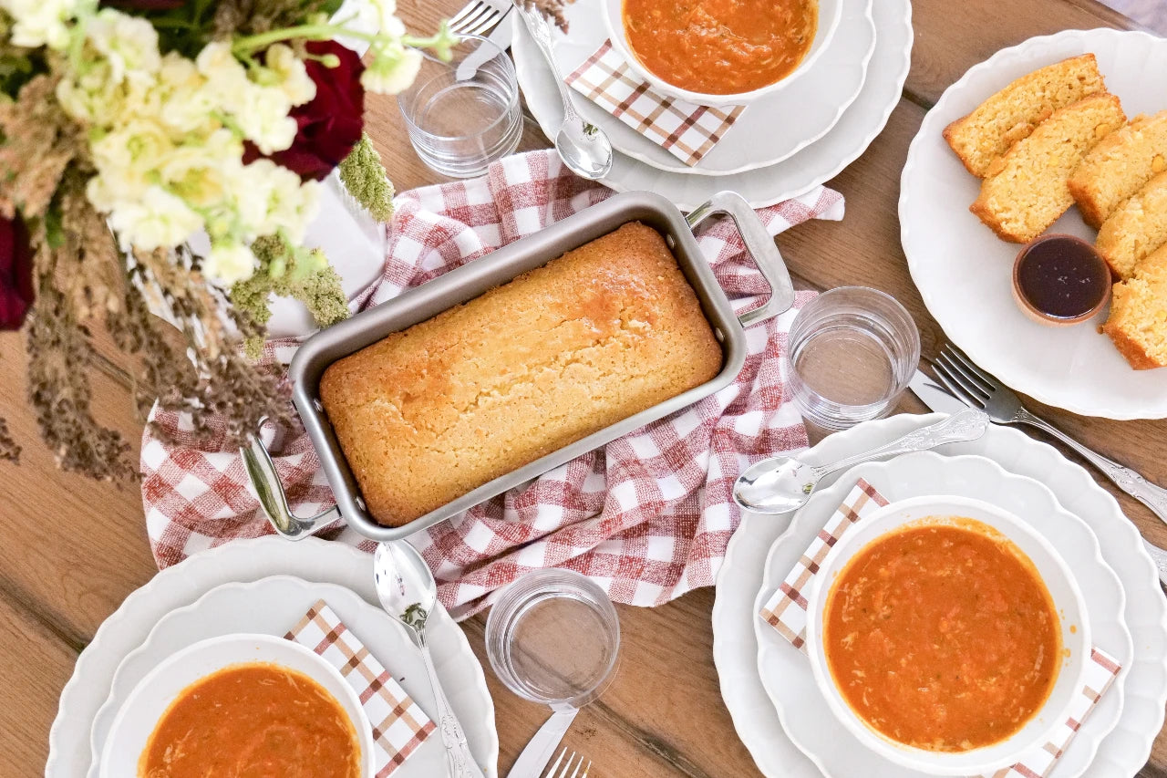 Stainless steel loaf pan with handles holding a baked loaf on a checkered cloth at a table with soup bowls and sliced bread