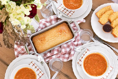Stainless steel loaf pan with handles holding a baked loaf on a checkered cloth at a table with soup bowls and sliced bread