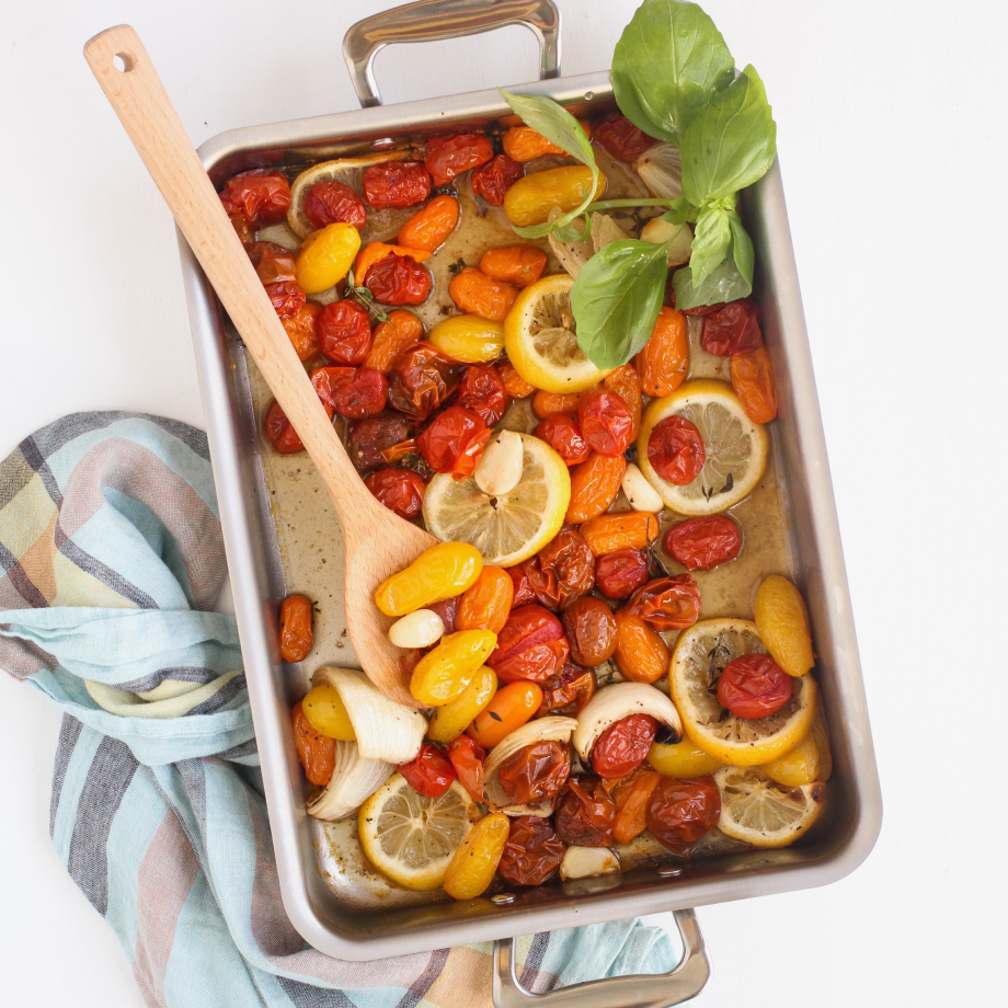 A stainless steel pan filled with roasted tomatoes, lemon slices, and fresh basil, with a wooden spoon resting on the side.