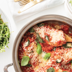 Saucepan of tomato-based pasta with melted cheese and basil, wooden spoon; bowls of arugula nearby.