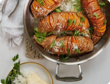Hasselback roasted potatoes in a stainless-steel pan garnished with grated Parmesan and parsley, dish of butter and napkin at the side