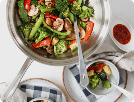 Top-down view of stir-fried vegetables in a stainless steel pan with bowls, fork, cloth napkins, and a small dish of chili sauce on a white surface