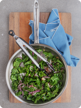 Pan with cooked kale and sautéed onions, metal tongs resting inside, on a wooden cutting board with a blue cloth napkin.