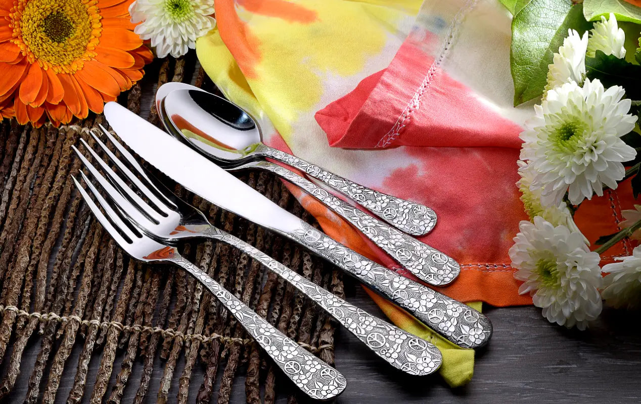 Top view of Woodstock flatware fork, knife, and spoon showcasing engraved floral handles on woven placemat with tie-dye napkin and flowers