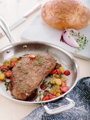 Cooked steak garnished with cherry tomatoes and herbs in a stainless steel pan, with a loaf of bread and sliced red onion on a cutting board in the background.