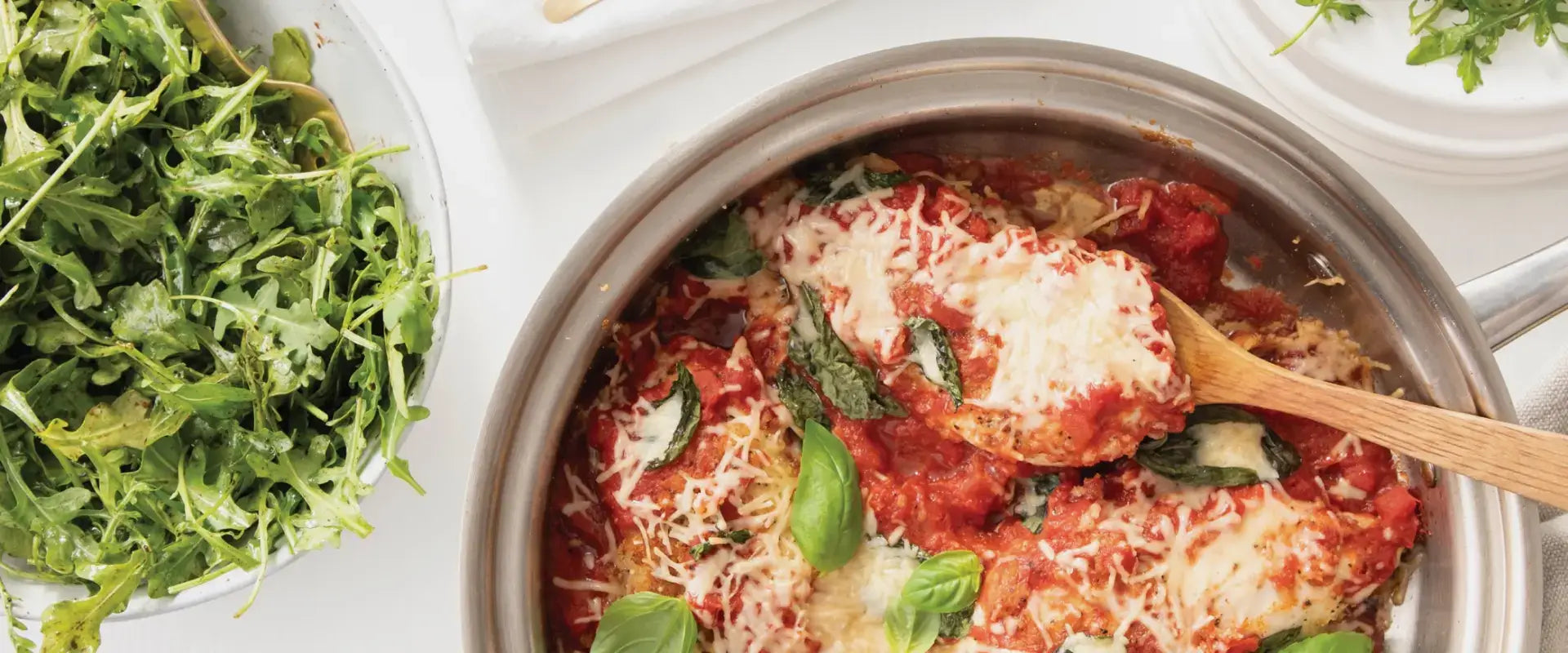 Skillet-baked chicken Parmesan topped with tomato sauce, melted cheese and basil, beside a bowl of arugula salad.