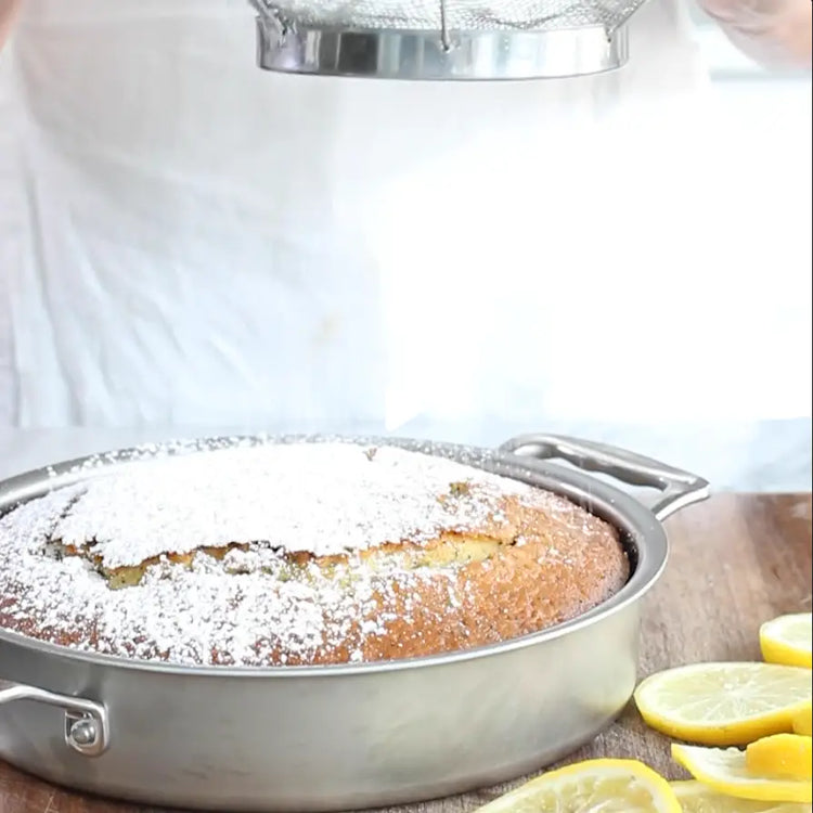Cake in a round metal pan with powdered sugar on top, surrounded by lemons.
